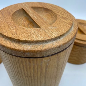 May include: Close-up of a set of wooden canisters with lids. The canisters are cylindrical with a light brown finish and a groove around the base of the lid. The lids have a carved-out handle. The wood grain is visible.