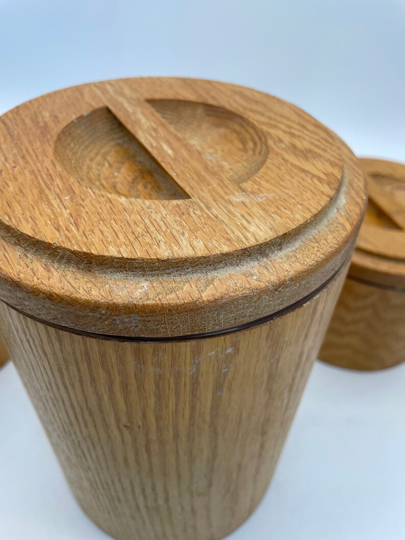 May include: Close-up of a set of wooden canisters with lids. The canisters are cylindrical with a light brown finish and a groove around the base of the lid. The lids have a carved-out handle. The wood grain is visible.