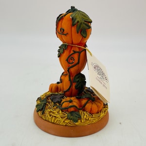May include: A ceramic cookie press featuring a stack of three orange pumpkins with green vines and leaves. The pumpkins sit on a bed of straw and small pumpkins. A tag reads "cookie press".