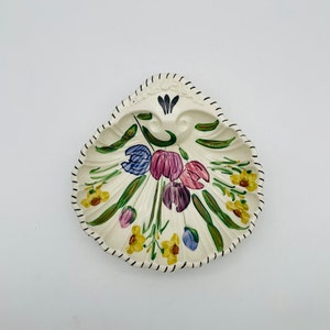 May include: A cream-colored, shell-shaped decorative dish with a hand-painted floral design. The flowers include purple, pink, blue, and yellow blooms with green leaves. The rim is outlined with a black, stitched pattern.