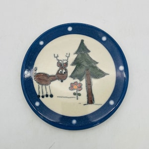 May include: A blue and white ceramic coaster with a hand-painted design of a deer, a pine tree, and a flower. The coaster has a white background with blue dots around the edge.