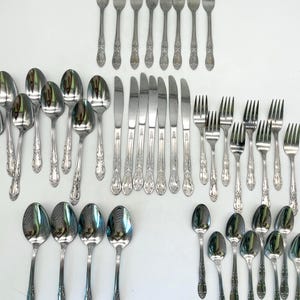 May include: A large collection of stainless steel flatware featuring a decorative floral pattern on the handles. The set includes various sizes of forks, spoons, and serrated knives, all arranged in organized rows against a plain white background.
