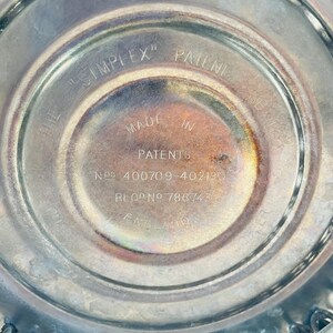 May include: A circular metallic base with engraved text reading THE SIMPLEX PATENT MADE IN ENGLAND PATENTS NOS 400709-402190 REG NO 786743. The surface has a polished finish with some tarnishing and a series of small metal loops along the outer edge.