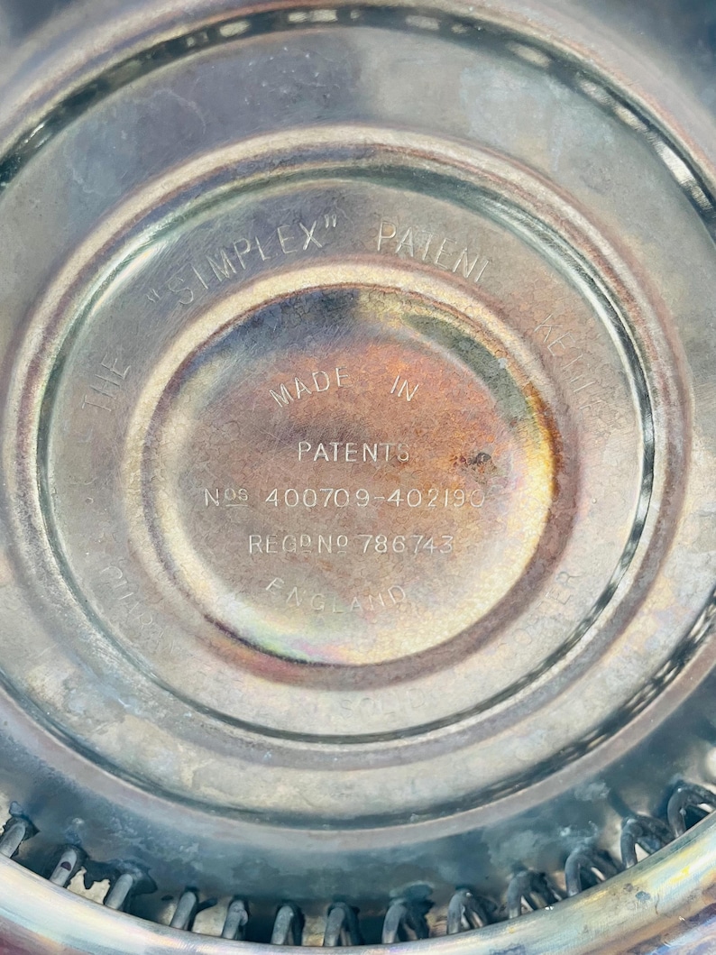May include: A circular metallic base with engraved text reading THE SIMPLEX PATENT MADE IN ENGLAND PATENTS NOS 400709-402190 REG NO 786743. The surface has a polished finish with some tarnishing and a series of small metal loops along the outer edge.