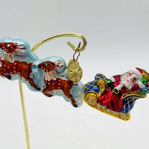May include: A Christmas ornament featuring Santa Claus in a sleigh pulled by two reindeer. The ornament is painted with bright colors and has a gold-colored charm hanging from it.