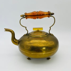 Może przedstawiać: Zabytkowy mosiężny czajnik o zaokrąglonym korpusie, zakrzywionej wylewce i uchwycie z ozdobnym uchwytem w kolorze bursztynu. Czajnik posiada małe nóżki i pokrywkę z pasującym bursztynowym akcentem.