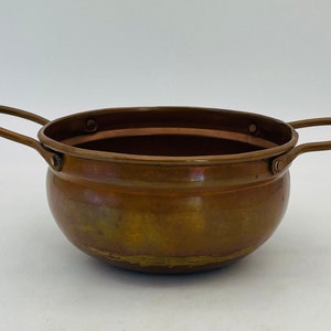 May include: A copper-coloured, round bowl with two curved handles. The bowl has a wide opening and a slightly rounded base. The copper has a warm, aged patina, with some areas showing a darker, more tarnished appearance. The handles are attached to the sides.