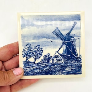 Può includere: Una piastrella quadrata in ceramica con un'illustrazione blu e bianca di un mulino a vento tradizionale accanto a una piccola casa e alberi. La scena raffigura un paesaggio rurale con un cielo nuvoloso, in un classico stile blu di Delft.