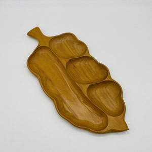 May include: A wooden serving tray in the shape of a leaf, featuring multiple compartments. The tray is made of light brown wood and has a smooth, polished finish. The design includes a handle and several sections for serving snacks or appetizers.