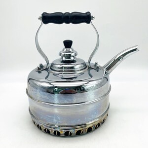 May include: A polished chrome tea kettle featuring a cylindrical body with a tiered base containing circular ventilation holes. The kettle has a black handle, a matching black knob on the removable lid, and a curved spout extending from the side.