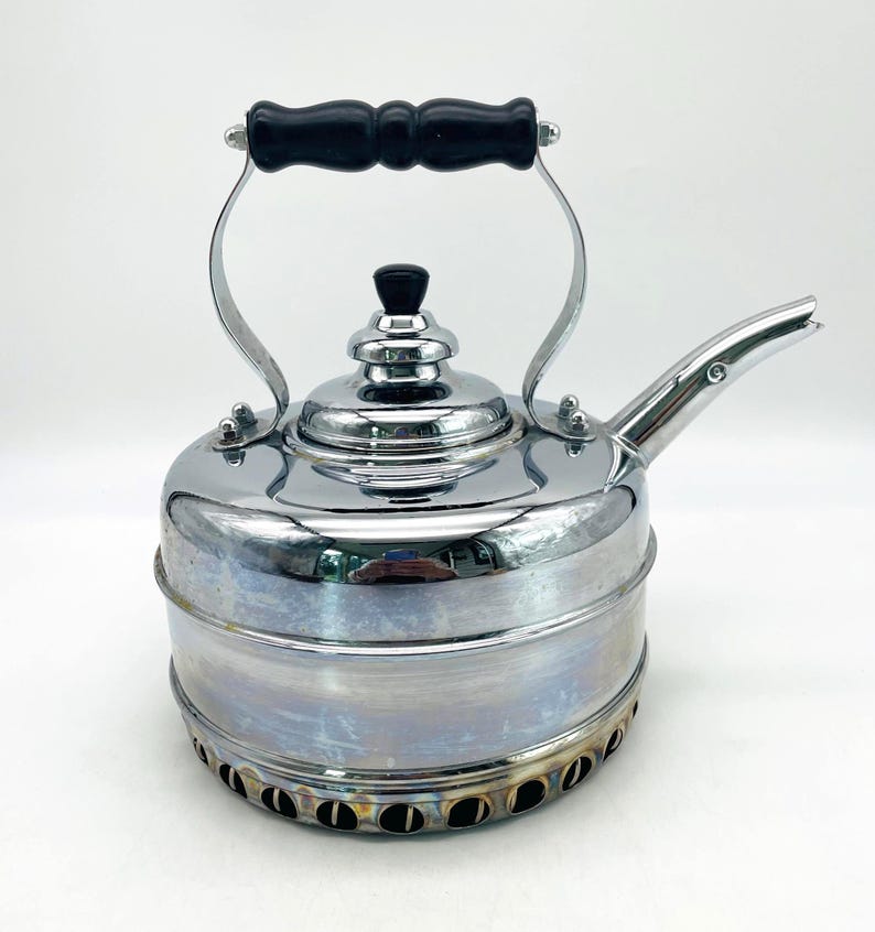 May include: A polished chrome tea kettle featuring a cylindrical body with a tiered base containing circular ventilation holes. The kettle has a black handle, a matching black knob on the removable lid, and a curved spout extending from the side.