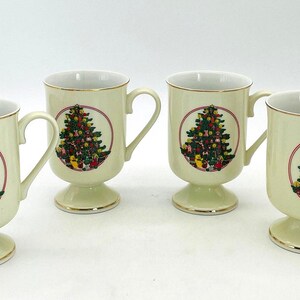 May include: A set of four cream-colored ceramic mugs, each featuring a pedestal base and a gold-trimmed rim. Every mug displays a circular graphic of a decorated Christmas tree with colorful ornaments and gifts at the base on the front side.