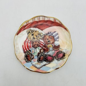 Peut inclure: Une petite assiette ronde en céramique avec un bord doré présente une illustration fantaisiste de trois ours en peluche en pyjama, profitant d'une scène de coucher paisible. Les ours tiennent des tasses et lisent un livre. L'assiette est décorée d'un motif à carreaux rouge, rose et bleu.