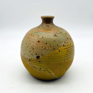 May include: A handcrafted ceramic vase with a rounded body and a narrow neck. The vase features a speckled, earthy color palette with shades of green, brown, and yellow. The lower portion is a vibrant yellow, adding a pop of color to the design.