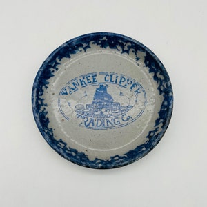 May include: A round, stoneware plate with a blue rim and a central design. The plate features the text "YANKEE CLIPPER TRADING CO." in blue, along with a ship illustration. The rim has a decorative blue pattern.