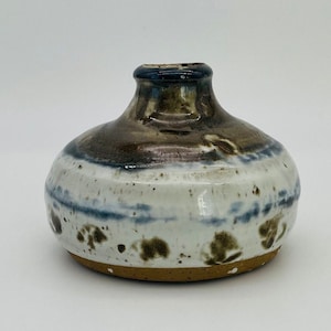 Può includere: Un piccolo vaso in ceramica fatto a mano con un corpo arrotondato e un collo stretto. Il vaso presenta una base bianca macchiettata, una fascia blu e marrone e una parte superiore marrone scuro. La base è di colore argilla naturale non smaltata.