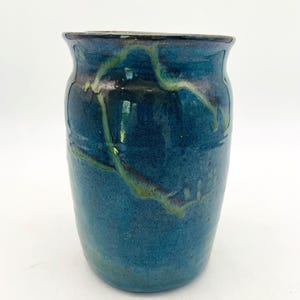 May include: A handcrafted ceramic vase with a dark blue glaze and streaks of green and yellow. The vase has a cylindrical shape with a slightly flared rim. The surface has a textured appearance, adding to its unique, artistic appeal.