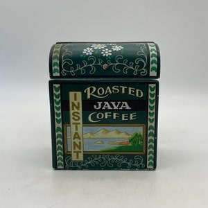 Puede incluir: Una lata de café de metal verde vintage. La parte frontal de la lata dice "Roasted Java Coffee" e "Instant". Presenta una escena de paisaje y diseños florales. La tapa arqueada está decorada con margaritas blancas. La lata mide unos 10 cm de alto.