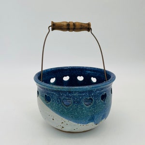 May include: A handcrafted ceramic basket with a blue and white glaze. The basket features heart-shaped cutouts around the rim and a wooden handle with a metal wire. The basket is approximately 6 inches in diameter and 5 inches tall.