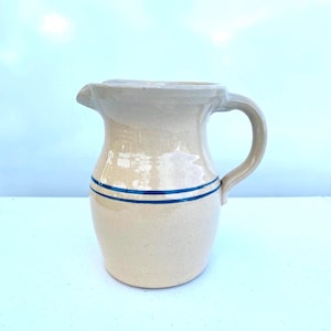 May include: A beige ceramic pitcher with a handle and spout. Two horizontal blue stripes encircle the body of the pitcher. The pitcher has a rustic, vintage appearance.