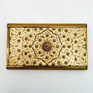 May include: Rectangular vintage compact case with a cream-colored floral design. The case has a gold-colored metal frame and a central medallion with red and gold accents. The floral pattern includes gold, red, and blue details.