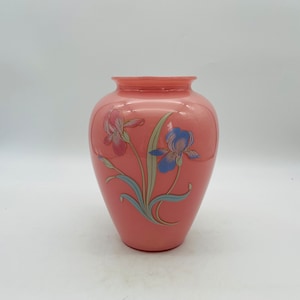May include: A peach-colored glass vase with a wide body and a narrow neck. The vase is decorated with two iris flower designs, one pink and one blue, with green and blue-gray leaves.