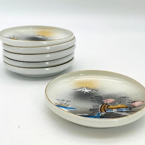 May include: A stack of six small, white ceramic dishes with a gold rim. One dish is separate, displaying a painted landscape with a mountain, trees, and a building. The dishes are likely for serving appetizers or small portions.