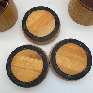 May include: Wooden coasters with a dark brown rubber ring and a light brown wooden centre. The coasters are round and have a natural wood grain pattern. Several wooden containers with a brown interior are also visible.