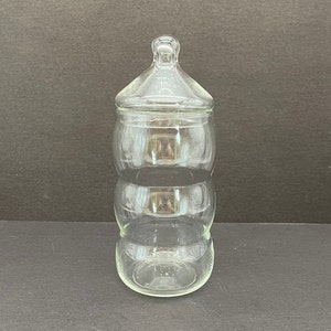 May include: A clear glass jar with a lid. The jar has a unique shape with three stacked, rounded sections. The lid has a small knob on top. The jar is empty and sits on a dark surface.