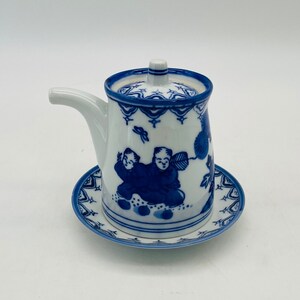 Könnte beinhalten: Eine weiße und blaue Keramik-Sauciere mit passender Untertasse. Die Sauciere hat einen Ausguss, einen Deckel und einen Griff. Das Design umfasst blaue Illustrationen von Figuren und floralen Mustern. Die Untertasse ist rund mit einem passenden blau-weißen Design.