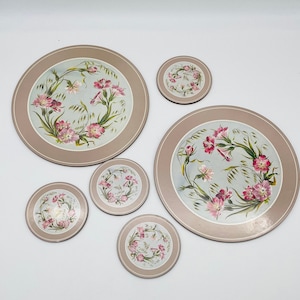 May include: A set of round placemats and coasters. The placemats and coasters have a light brown border and a central design of pink flowers and green leaves on a light blue background. The set includes two large placemats and five coasters.
