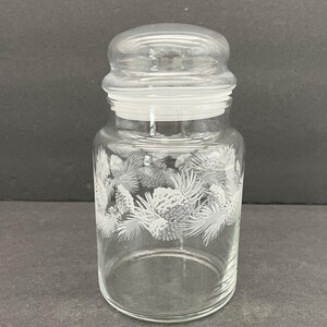 Peut inclure: Un bocal en verre transparent avec un couvercle, orné d'un motif de pommes de pin et d'aiguilles blanches. Le bocal est cylindrique avec un couvercle arrondi. Le bocal est vide.