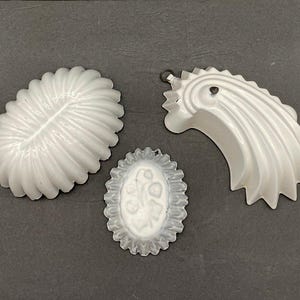May include: Three white, vintage-style molds for baking or crafting. The molds include an oval shape with a floral design, a shell-shaped mold, and a decorative, curved mold with a hanging loop.