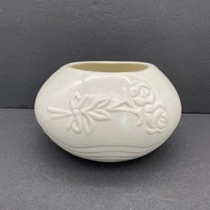 May include: A cream-colored, oval-shaped ceramic vase. The vase features a raised floral design of roses and a bow. The vase has a smooth, glossy finish and a wide opening. The vase is a decorative item.