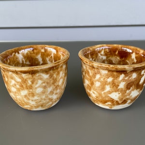May include: Two small, handmade ceramic bowls with a speckled brown and cream pattern. The bowls have a rounded shape and a glossy finish, perfect for serving dips or small portions.
