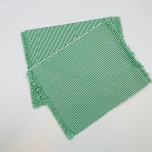 May include: Two square, light green placemats with a striped pattern and fringed edges. The placemats have a white border and are made of a textured fabric. The placemats are designed for dining and home decor.