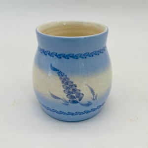 Puede incluir: Una taza de cerámica con cuerpo redondeado que presenta un patrón de rayas horizontales en azul claro y crema. La taza está decorada con un motivo pintado en azul oscuro de trigo o flores en el centro y dos bandas horizontales de hojas en relieve.