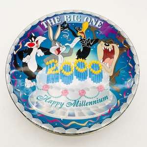 May include: A round, metal tin featuring Looney Tunes characters Sylvester, Bugs Bunny, Daffy Duck, and Taz. The tin has a blue background with a cake that reads "2000" and "Happy Millennium". The top of the tin reads "The Big One".
