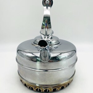 May include: A polished chrome stovetop kettle with a cylindrical body and a curved spout. The kettle features a vertical handle with a black circular grip at the top. The base of the kettle has a series of circular cutouts and shows heat discoloration.