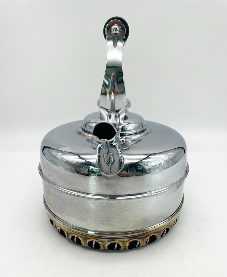 May include: A polished chrome stovetop kettle with a cylindrical body and a curved spout. The kettle features a vertical handle with a black circular grip at the top. The base of the kettle has a series of circular cutouts and shows heat discoloration.