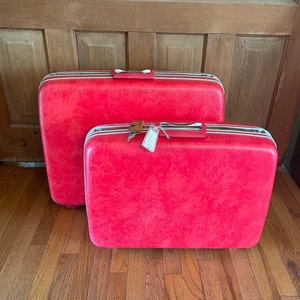 May include: Two vintage red suitcases with white and silver trim, one larger than the other. The suitcases have handles and are sitting on a wooden floor. One suitcase has a luggage tag attached.
