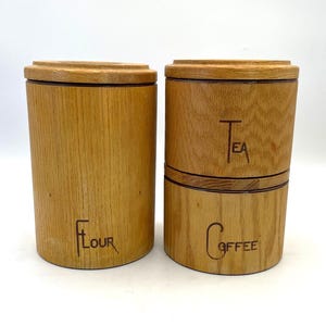 May include: Two wooden canisters with lids, one labeled "Flour" and the other labeled "Tea" and "Coffee". The canisters are cylindrical and made of light-colored wood. The text is in a simple, elegant font.