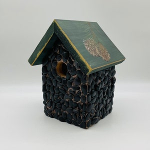May include: A decorative birdhouse with a dark green roof and a body covered in small, black stones. The roof has a painted pine cone design. The birdhouse has a round entry hole and is designed to attract birds.