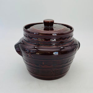 May include: A dark brown ceramic casserole dish with a lid. The dish features two handles and decorative floral and wave patterns around the sides. The lid has a small knob and a similar pattern. The dish is glazed and has a glossy finish.