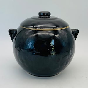 May include: A black ceramic lidded pot with two side handles. The pot has a glossy finish and a gold-colored trim around the lid. The lid has a small knob on top. The pot sits on a white surface.