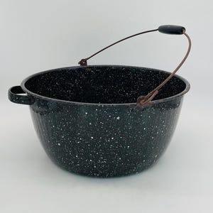 May include: A black speckled enamelware pot with a metal handle and side handle. The pot has a rounded shape and a dark speckled pattern. The handle is made of metal and has a wooden grip. The pot is likely used for cooking or storage.