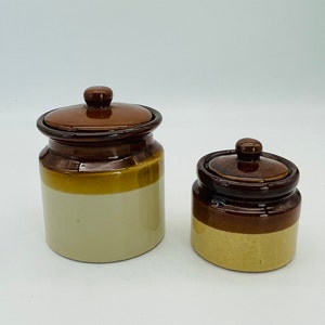 May include: Two ceramic jars with lids. The jars are brown, tan, and cream-colored. The larger jar is taller than the smaller jar. The lids have a small knob on top. The jars are likely for kitchen storage.