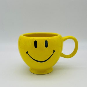 May include: A bright yellow ceramic mug with a smiling face design. The mug has a rounded shape, a handle, and a small base. The smiley face features black eyes and a curved black mouth.