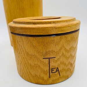 May include: Wooden tea canister with a lid and a matching tall container in the background. The canister is light brown with a dark brown band and the word "TEA" printed on the front. The wood grain is visible.