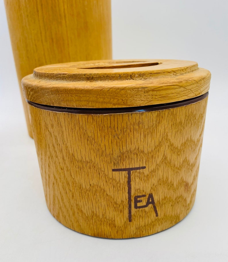 May include: Wooden tea canister with a lid and a matching tall container in the background. The canister is light brown with a dark brown band and the word "TEA" printed on the front. The wood grain is visible.